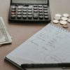 An open notebook, a pen, a calculator and cash on a kitchen table.