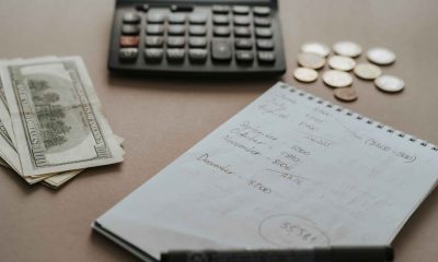 An open notebook, a pen, a calculator and cash on a kitchen table.