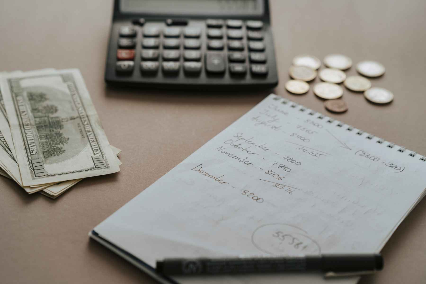 An open notebook, a pen, a calculator and cash on a kitchen table.